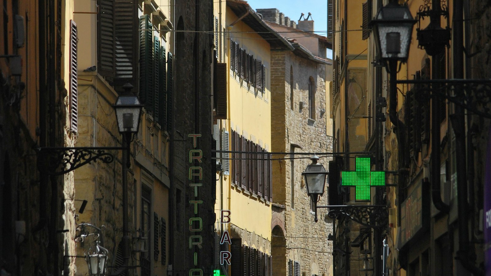 A green pharmacy cross sign on a narrow historic street in Italy